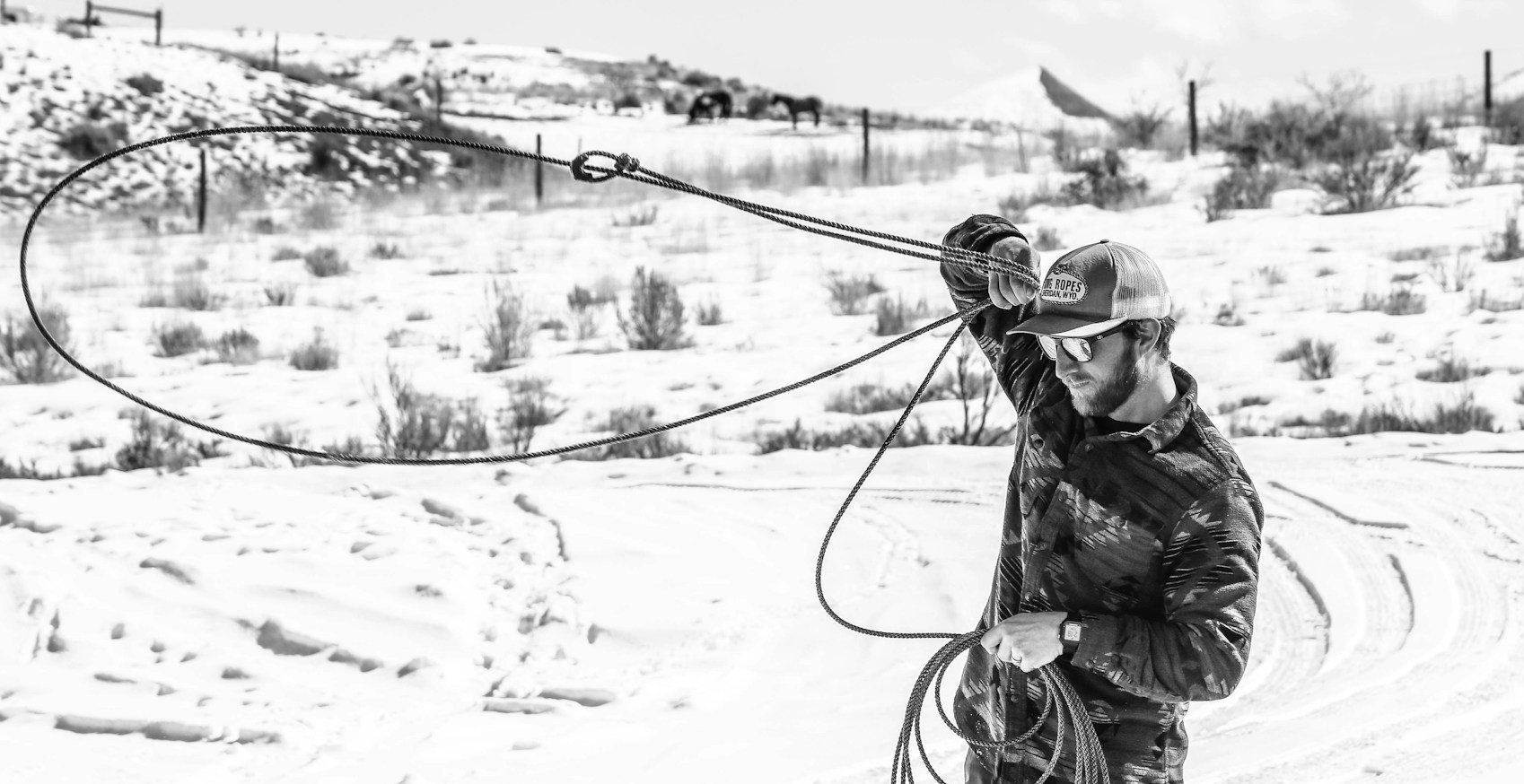 A man swings a lasso in a dry desert or mountain scene. The image is in black and white so it looks as if the ground is snowy.