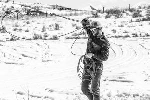 A black and white photo of a man swinging a lasso. The landscape behind him is mostly white and dry.