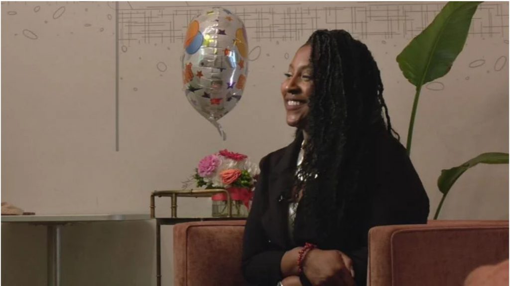 JaNae Martin, a black woman in a black blazer, sits in a chair, smiling. A birthday balloon floats behind her.