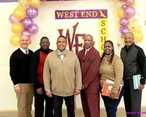 A group of people standing next to each other in front of a small balloon arch with the words "West End School" painted on the wall behind them.