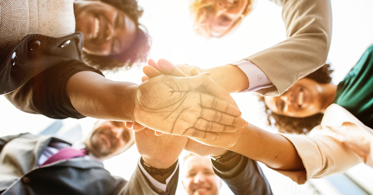 A group of people stand in a circle and stack their hands. The picture is taken from below the stack, so you can see their smiling faces and the flare of the sun.