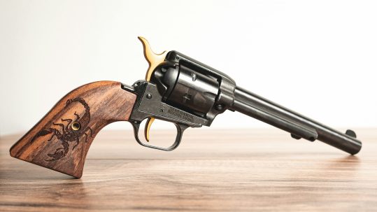 A revolver is balanced on its handle and barrel on a wood surface. The handle is wooden with a scorpion burnished into the wood. The rest of the gun is gray metal.