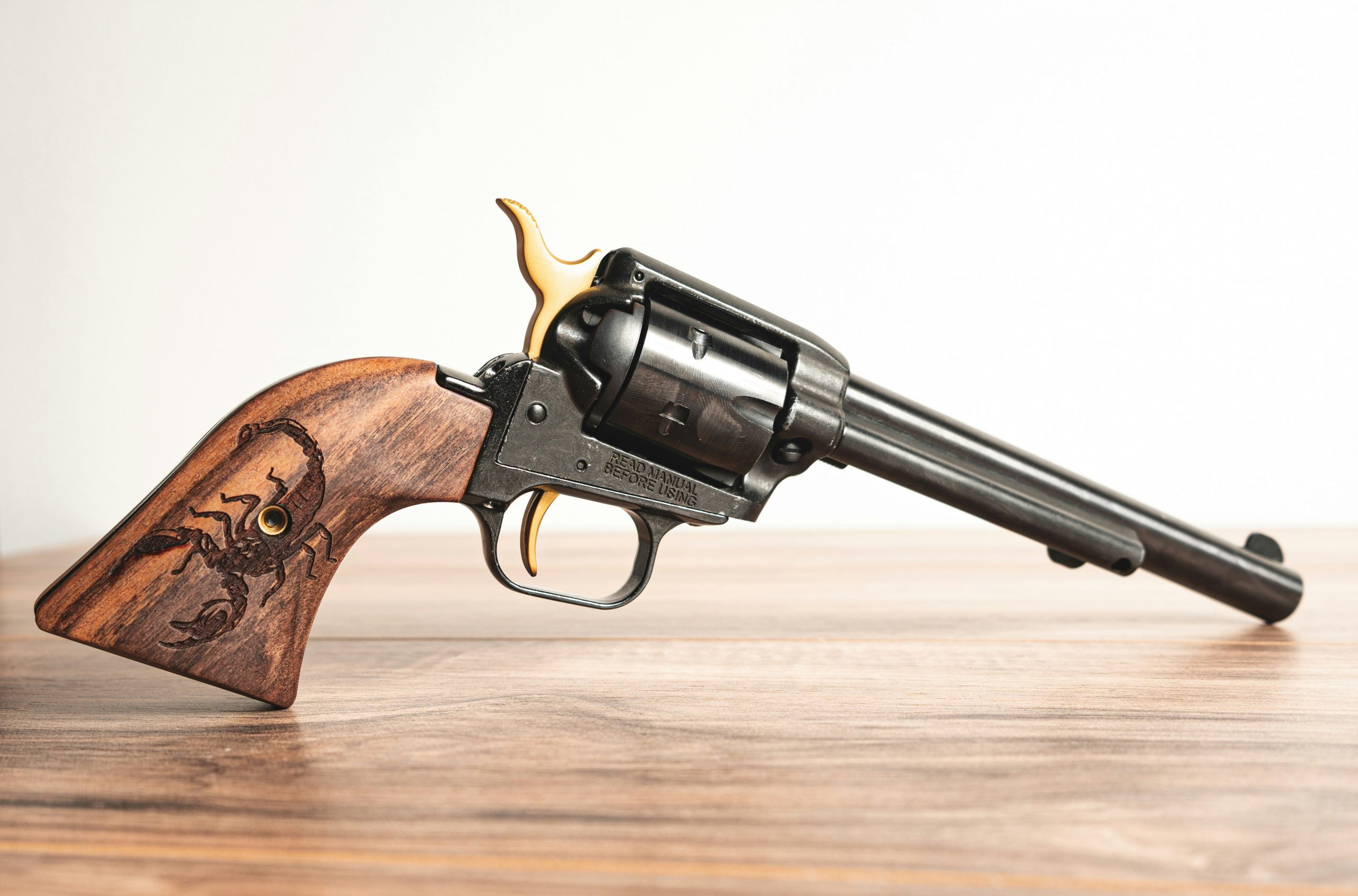 A revolver is balanced on its handle and barrel on a wood surface. The handle is wooden with a scorpion burnished into the wood. The rest of the gun is gray metal.