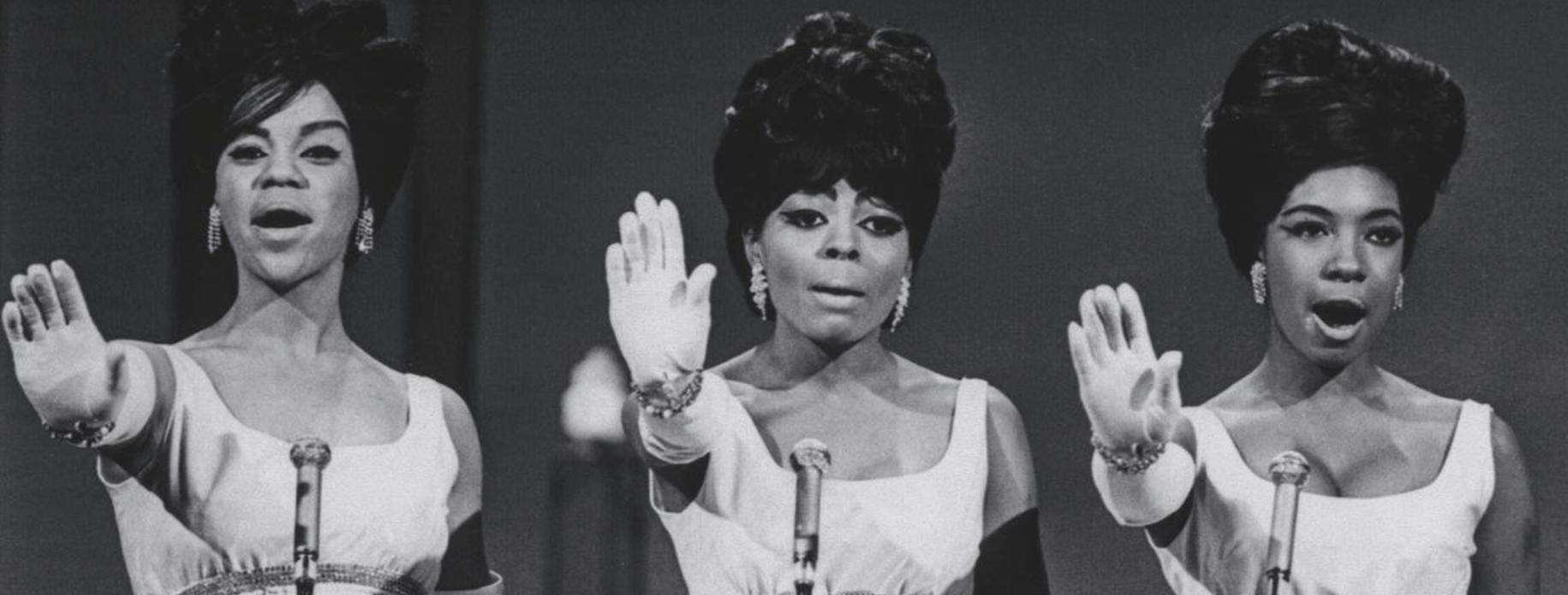 The Supremes (three Black women) singing into mics with their hands out in front of them.