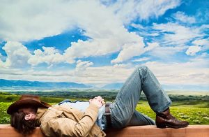 man laying down under a blue sky with a cowboy hat on his face and one leg bent