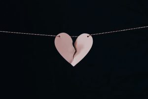 A heart shaped piece of paper is hung on a thin string in front of a black background. The paper is torn down the middle in a broken heart zig zag.