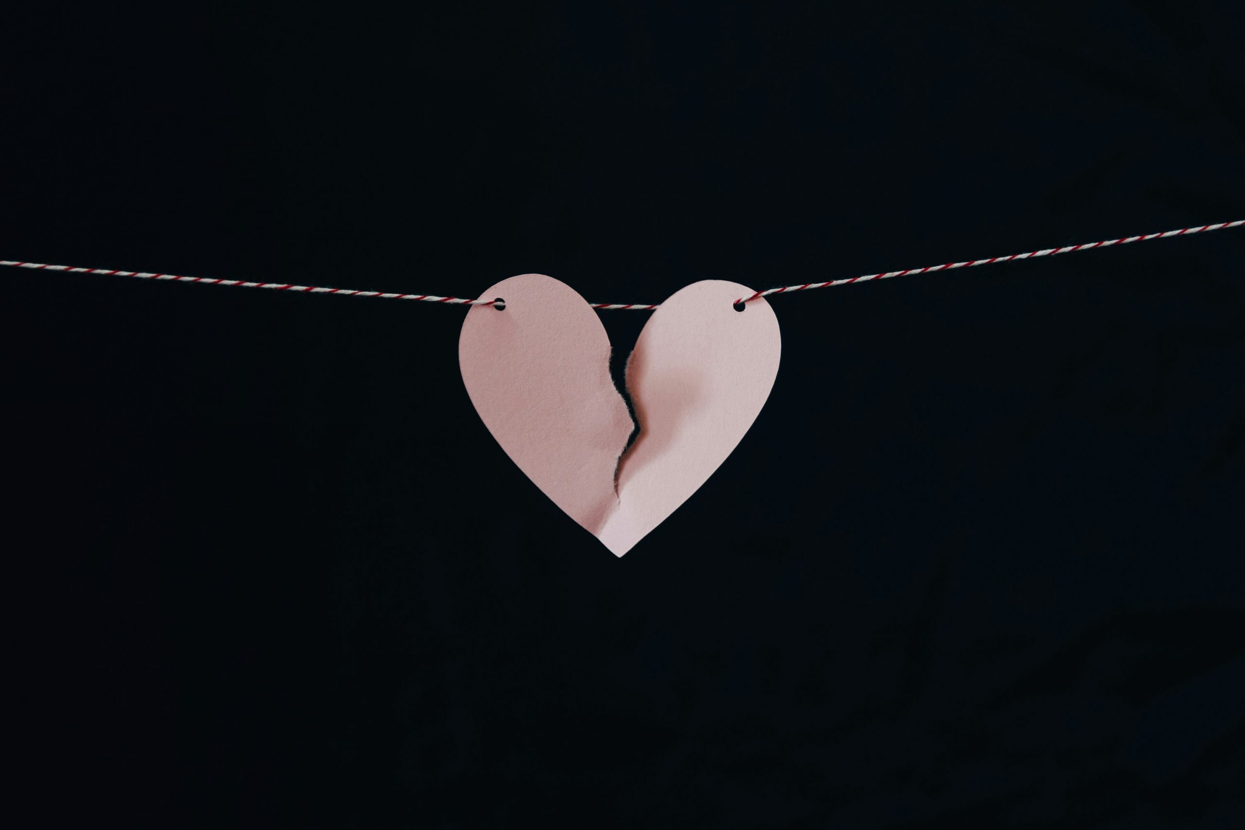 A heart shaped piece of paper is hung on a thin string in front of a black background. The paper is torn down the middle in a broken heart zig zag.
