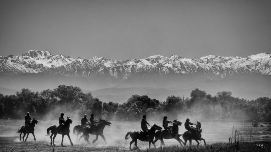 A black and white photo of a group of people riding horses. In the background, a mountain range is visible through fog.